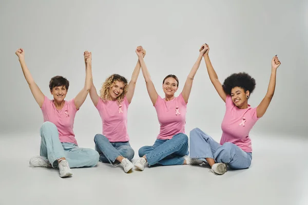 Excitado interracial mujeres diferentes edad cogidas de la mano en gris telón de fondo, conciencia de cáncer de mama - foto de stock