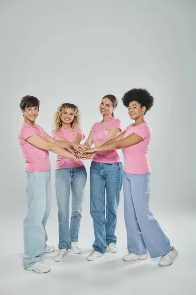 Glückliche interrassische Frauen unterschiedlichen Alters, die vor grauem Hintergrund Hände sammeln, Bewusstsein für Brustkrebs — Stockfoto