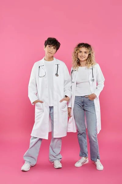 Весёлые женщины-врачи в белых халатах, стоящие на розовом фоне, радость, осознание рака молочной железы, женщины — стоковое фото
