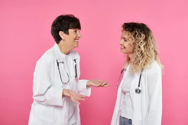 Happy female doctors in white coats chatting on pink backdrop, joy, breast cancer awareness, women — Stock Photo