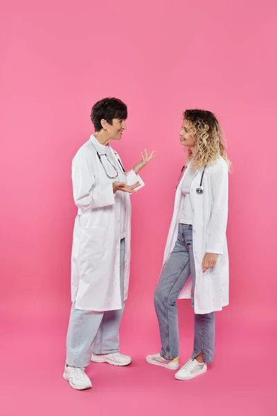 Heureuses femmes médecins en manteaux blancs bavardant sur fond rose, sensibilisation au cancer du sein, femmes — Photo de stock