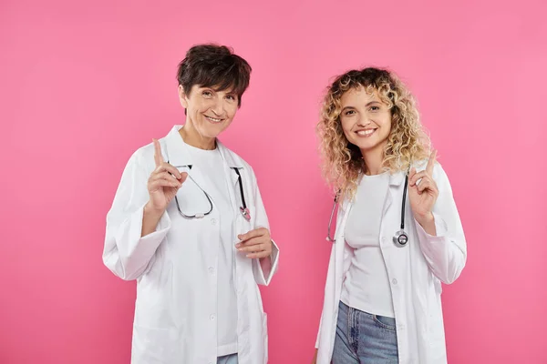 Female doctors in white coats warning on pink backdrop, smile, breast cancer awareness, women — Stock Photo