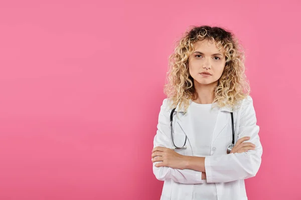 Концепция информированности о раке молочной железы, кудрявая врач смотрит в камеру, розовый фон, женщина — стоковое фото