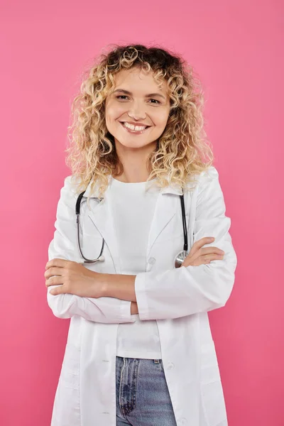 Концепция информированности о раке молочной железы, кудрявая врач смотрит на камеру, розовый фон, улыбка, женщина — стоковое фото