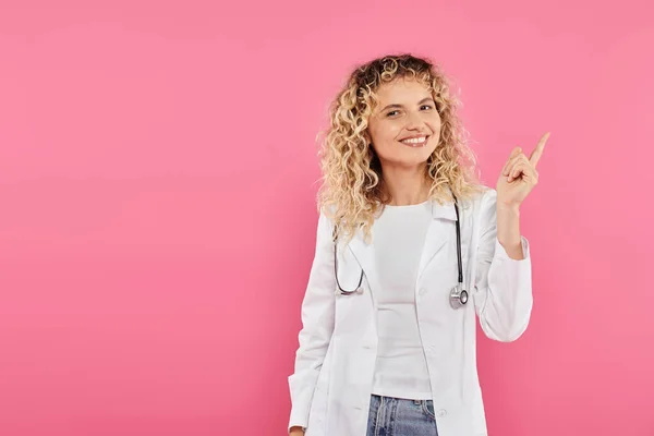 Концепция информированности о раке молочной железы, счастливая женщина врач указывая вверх, розовый фон, улыбка, женщина — стоковое фото