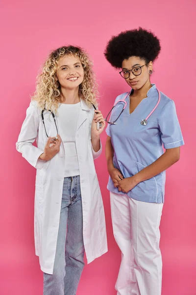 Онкологи, міжрасові жінки-лікарі в білих пальто на рожевому тлі, обізнаність про рак молочної залози — стокове фото