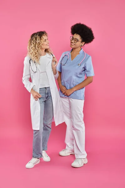 Oncologues féminines, médecins interraciaux en blouse blanche sur fond rose, sensibilisation au cancer du sein — Photo de stock