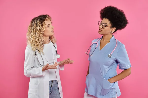 Oncologists chatting, interracial doctors in white coats on pink backdrop, breast cancer awareness — Stock Photo