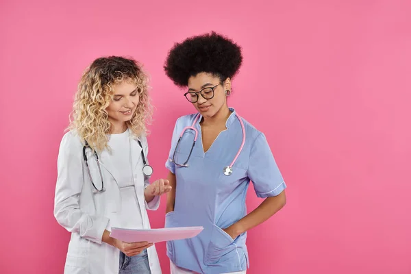 Oncologists discussing diagnosis, interracial doctors on pink backdrop, breast cancer awareness — Stock Photo