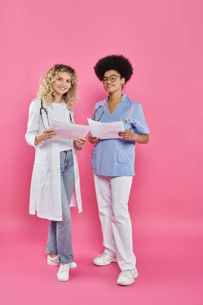 Oncologi femminili allegri, medici interrazziali su sfondo rosa, consapevolezza del cancro al seno — Foto stock