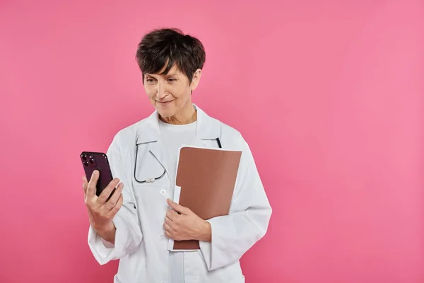 Зрелый онколог, женщина врач держит папку, используя смартфон, концепция рака молочной железы осведомленности — стоковое фото
