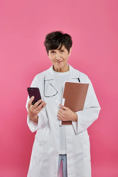 Oncologista maduro, médico feminino segurando pasta e smartphone, consciência do câncer de mama, idade em tecnologia — Fotografia de Stock