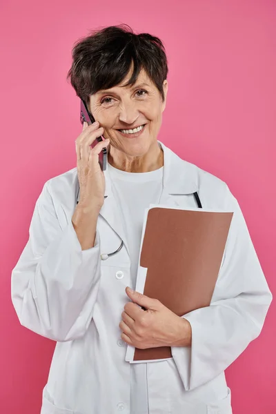 Oncólogo maduro, médico femenino sosteniendo la carpeta y hablando en el teléfono inteligente, conciencia del cáncer de mama - foto de stock