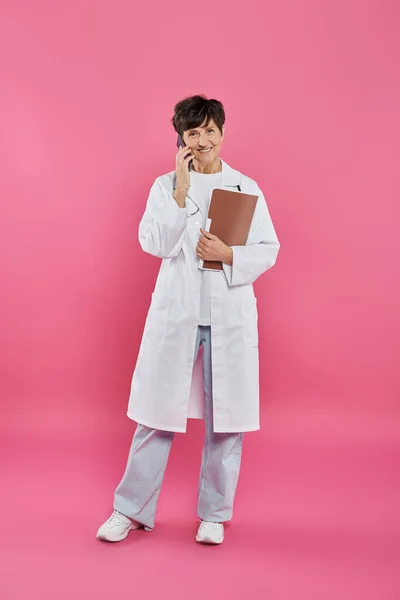 Oncólogo femenino, médico maduro sosteniendo la carpeta y hablando en el teléfono inteligente, conciencia del cáncer de mama - foto de stock