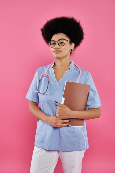 Breast cancer awareness, african american doctor, female oncologist with folder, pink backdrop — Stock Photo