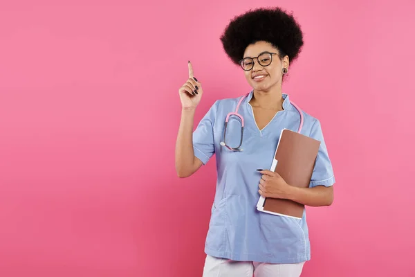 Conciencia del cáncer de mama, médico afroamericano, oncólogo feliz con carpeta, apuntando hacia arriba, rosa - foto de stock