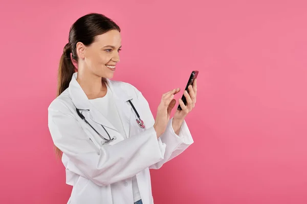 Médecin femme souriante en manteau blanc en utilisant smartphone isolé sur rose, concept de cancer du sein — Photo de stock