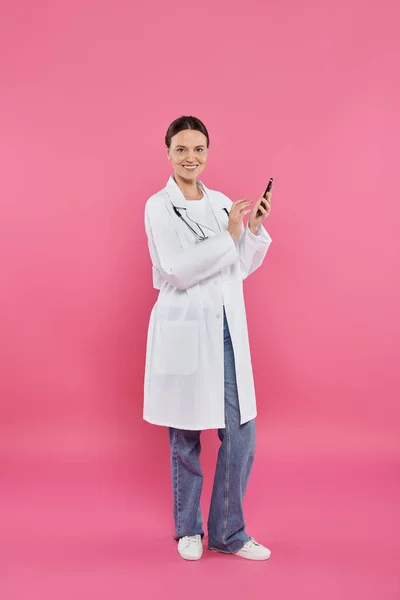 Sonriente doctor en blanco abrigo celebración de teléfono inteligente y mirando a la cámara en rosa, concepto de cáncer de mama - foto de stock