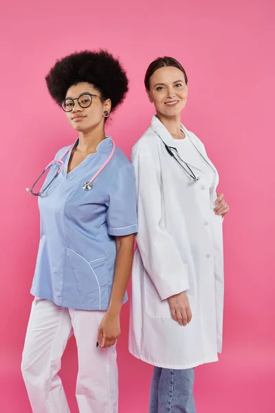 Усміхнені багатоетнічні лікарі дивляться на камеру ізольовані на рожевому, концепція раку молочної залози — стокове фото