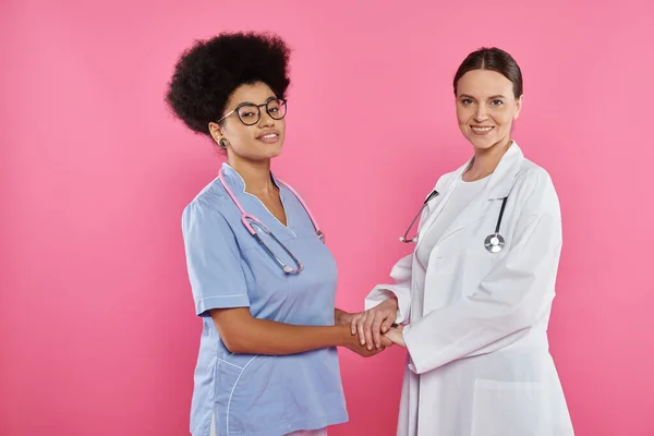 Smiling multiethnic doctors holding hands and standing isolated on pink, breast cancer awareness — Stock Photo
