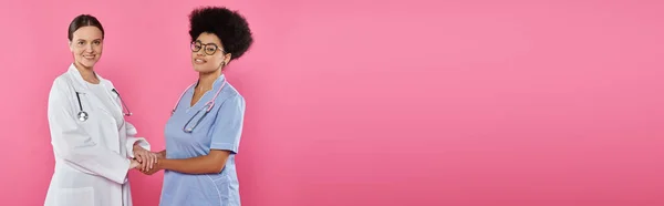 Médecins multiethniques positifs tenant les mains isolées sur rose, bannière, concept de cancer du sein — Photo de stock