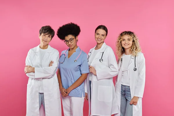 Médecins gais et multiethniques souriant à la caméra isolé sur rose, concept de cancer du sein — Photo de stock