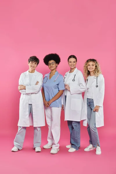 Full length of smiling interracial doctors standing together on pink, breast cancer month concept — Stock Photo
