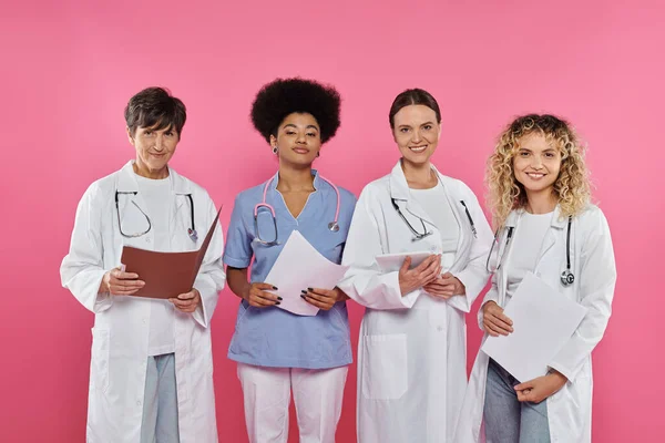 Médecins souriants multiethniques tenant des dossiers en papier et tablette numérique isolés sur rose, cancer du sein — Photo de stock