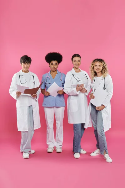 Souriant interracial médecins tenant des dossiers en papier et tablette numérique sur rose, cancer du sein mois — Photo de stock