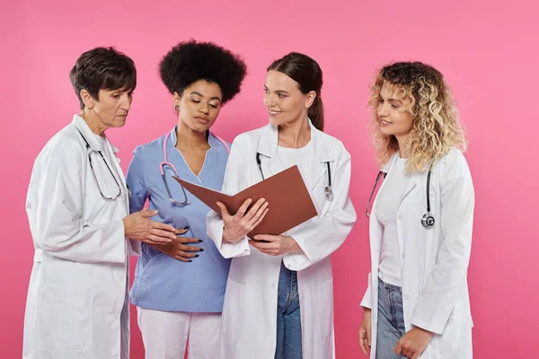 Усміхнений лікар тримає паперову теку біля багатоетнічних колег ізольовано на рожевому раку молочної залози — стокове фото