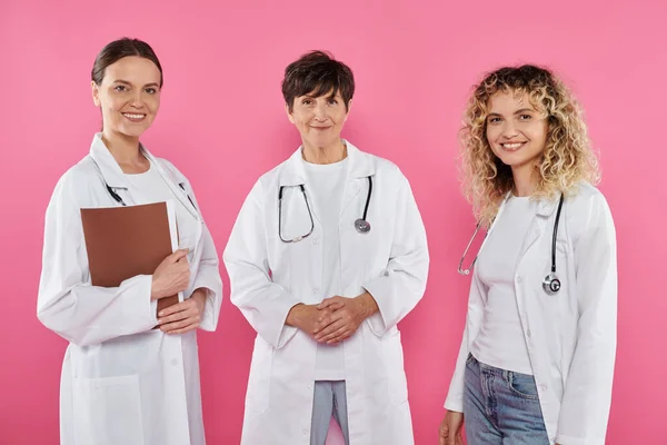 Lächelnde Ärztinnen in weißen Mänteln, die in die Kamera schauen, isoliert auf rosa, Brustkrebs-Bewusstsein — Stockfoto