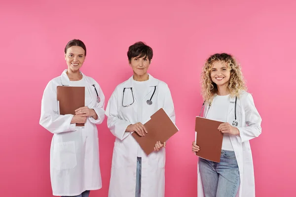 Fröhliche Ärzte halten Papiermappen in der Hand, während sie isoliert auf rosa, Brustkrebs-Konzept stehen — Stockfoto