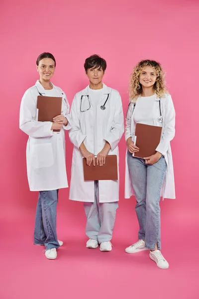 Heureuses femmes médecins tenant des dossiers en papier et debout sur fond rose, sensibilisation au cancer du sein — Photo de stock