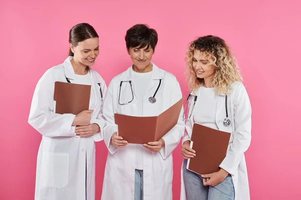 Усміхнені лікарі дивляться на паперову папку разом ізольовані на рожевому, місяць обізнаності про рак молочної залози — стокове фото