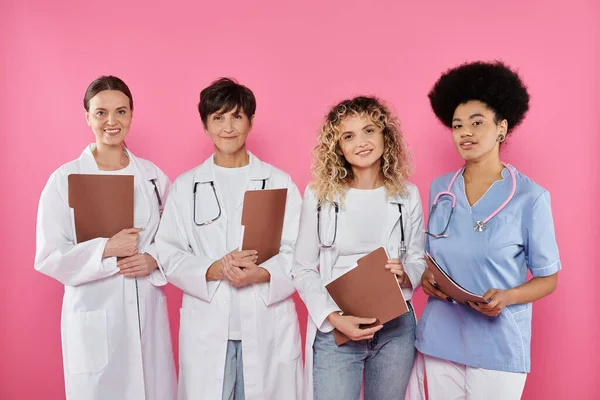 Happy interracial oncologists with paper folders looking at camera isolated on pink, breast cancer — Stock Photo