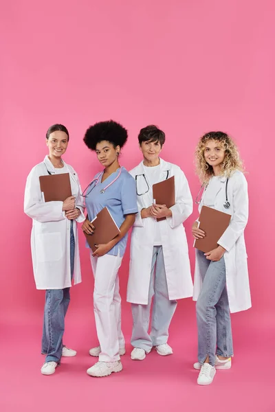 Улыбающиеся мультиэтнические врачи с бумажными папками, стоящими вместе на розовом, рак груди осведомленности — стоковое фото
