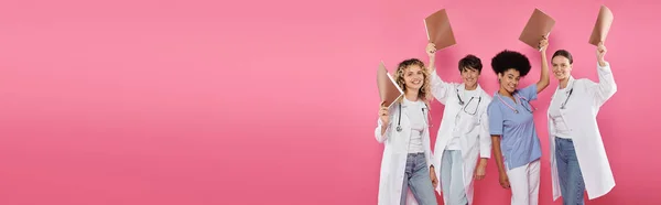 Sonrientes médicos multiétnicos sosteniendo carpetas de papel aisladas en rosa, pancarta, mes del cáncer de mama - foto de stock