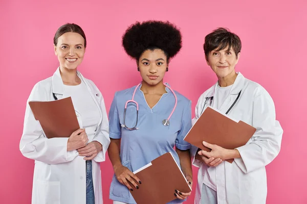 Sorridenti medici interrazziali in camici bianchi che tengono cartelle di carta isolate su rosa, cancro al seno — Foto stock