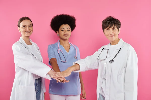 Positive multiethnic oncologists holding hands isolated on pink, breast cancer awareness — Stock Photo