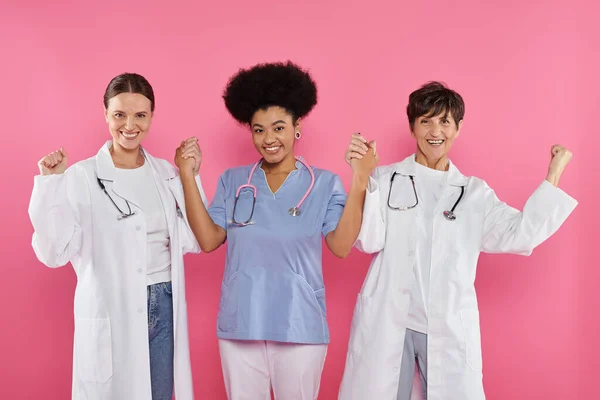 Excited multiethnic doctors oncologists holding hands isolated on pink, breast cancer month — Stock Photo