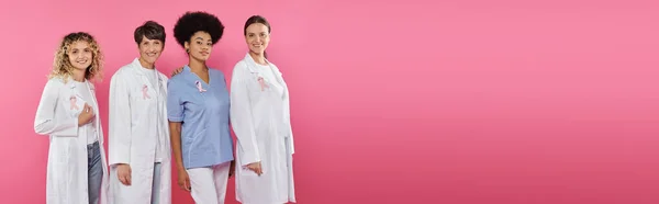 Médicos multiétnicos sonrientes con cintas en abrigos de pie aislados en rosa, pancarta, cáncer de mama - foto de stock