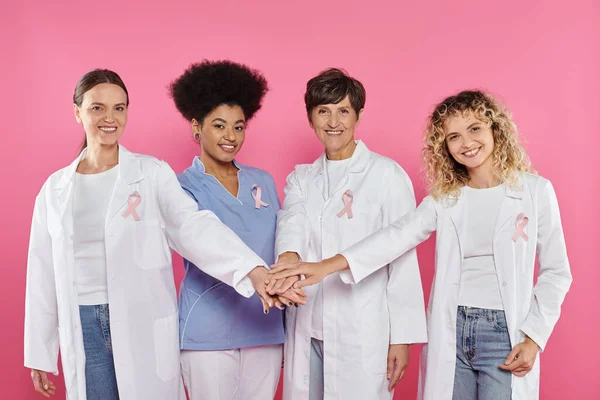 Улыбающиеся многонациональные врачи с лентами, держащиеся за руки, изолированные от розового, концепция рака груди — стоковое фото