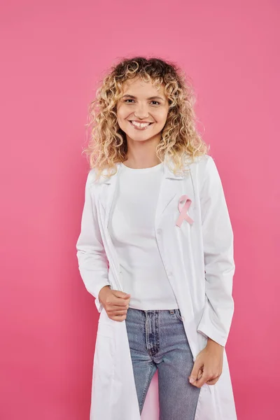 Medico sorridente con nastro su camice bianco isolato su rosa, concetto di cancro al seno — Foto stock