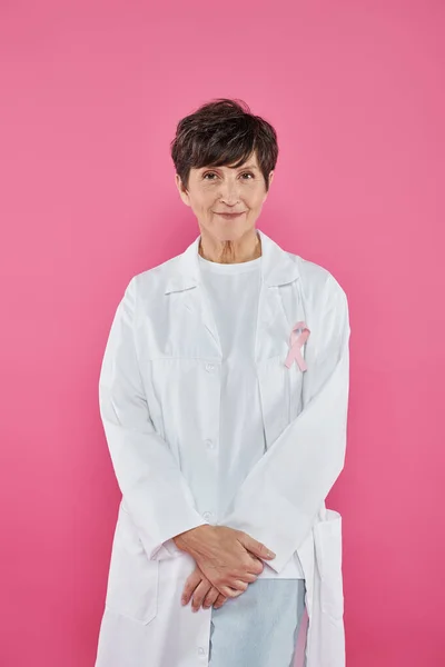 Brunette doctor with ribbon on white coat standing isolated on pink, breast cancer concept — Stock Photo