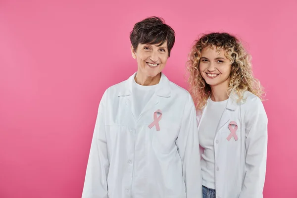Весёлые онкологи с лентами на белых халатах, изолированные от розового, концепция рака молочной железы — стоковое фото
