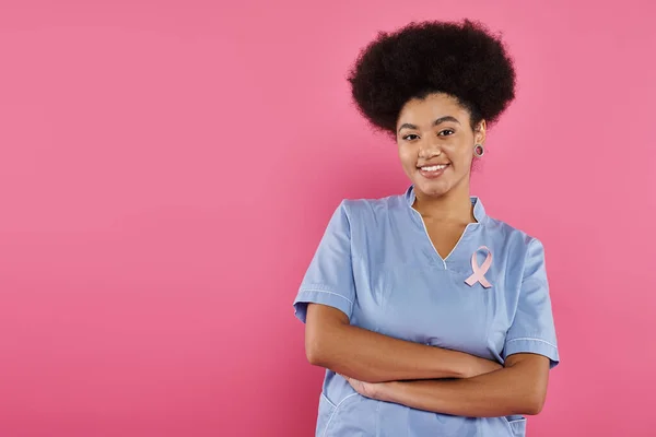 Médecin afro-américain souriant avec ruban traversant les bras isolé sur rose, concept de cancer du sein — Photo de stock