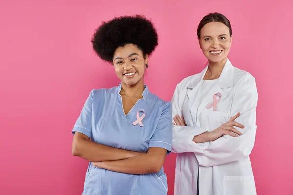 Радостные межрасовые онкологи с лентами, скрещивающими руки, изолированными на розовом месяце рака груди — стоковое фото