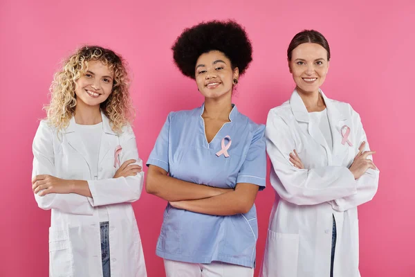 Interracial doctors with ribbons crossing arms and smiling on pink, breast cancer concept — Stock Photo