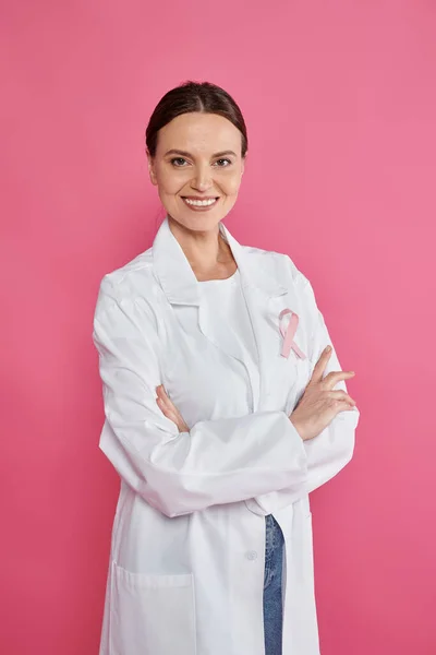 Femme oncologue joyeuse avec ruban croisant les bras isolé sur rose, mois du cancer du sein — Photo de stock