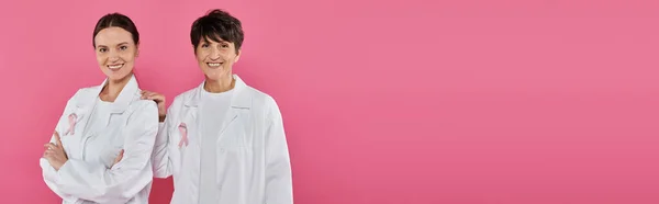 Medici sorridenti con nastri su camici bianchi in posa isolati su rosa, concetto di cancro al seno, striscione — Foto stock